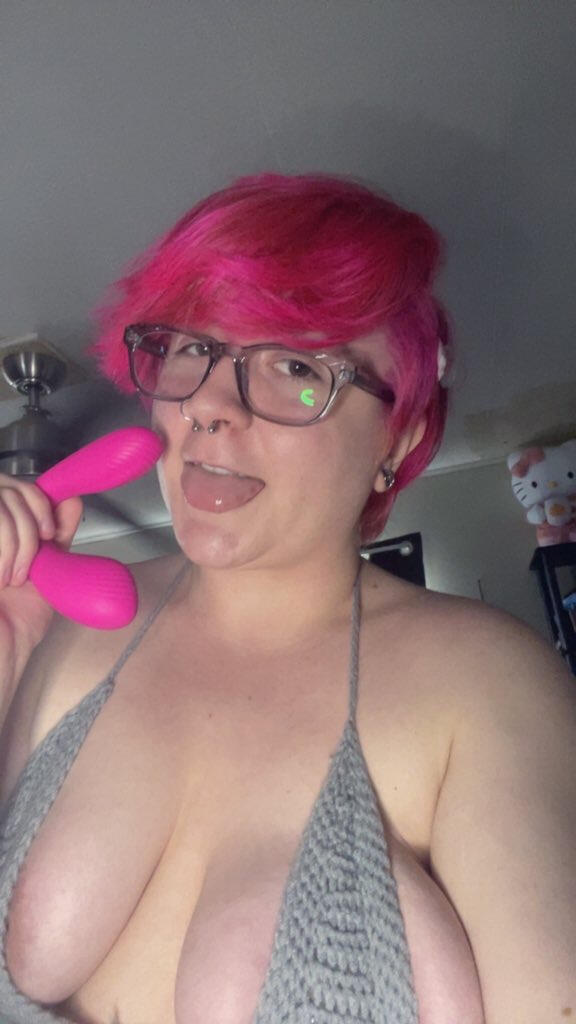 Image of Onyx, an adult with short pink hair and square glasses. He is posed holding a pink toy and sticking his tongue out.